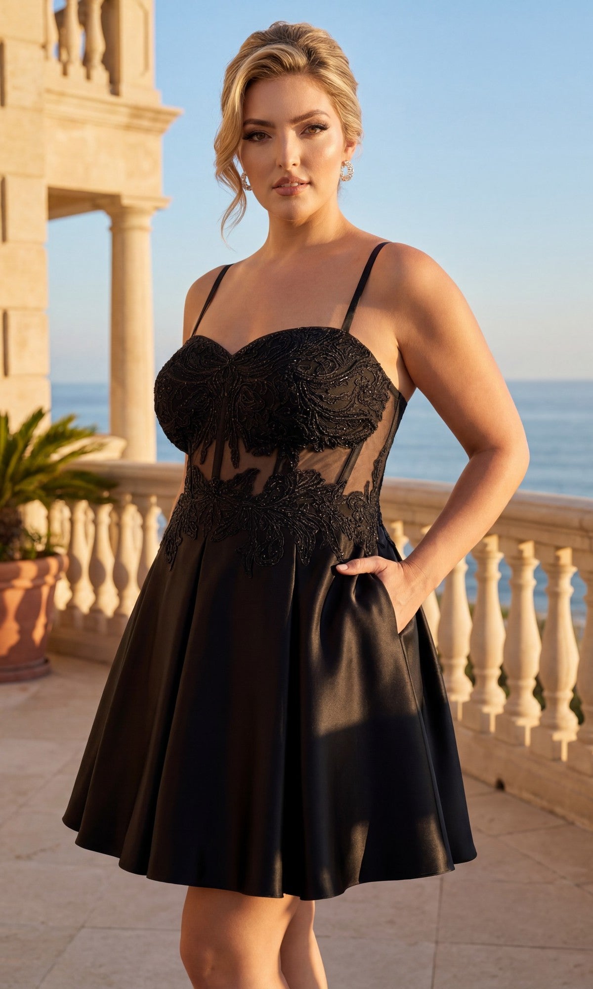 A woman wearing the Plus-Size Black Hoco Dress: Blondie Nites 6346BNW stands on a terrace overlooking the sea, with lace details on her short black dress and stone railings and columns in the background.